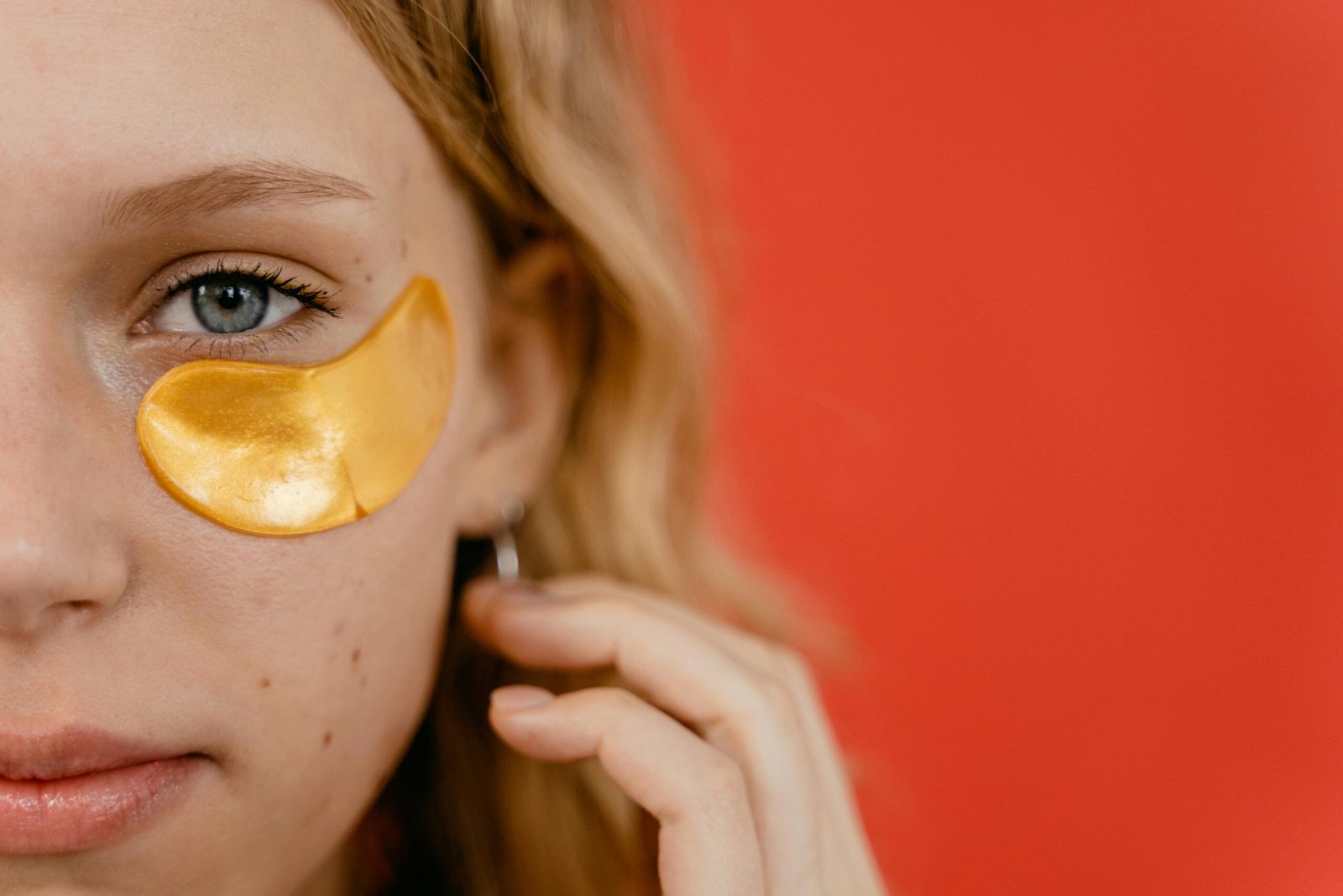 Close-up of a young woman with a gold eye patch against a vibrant red background, highlighting skincare routine.