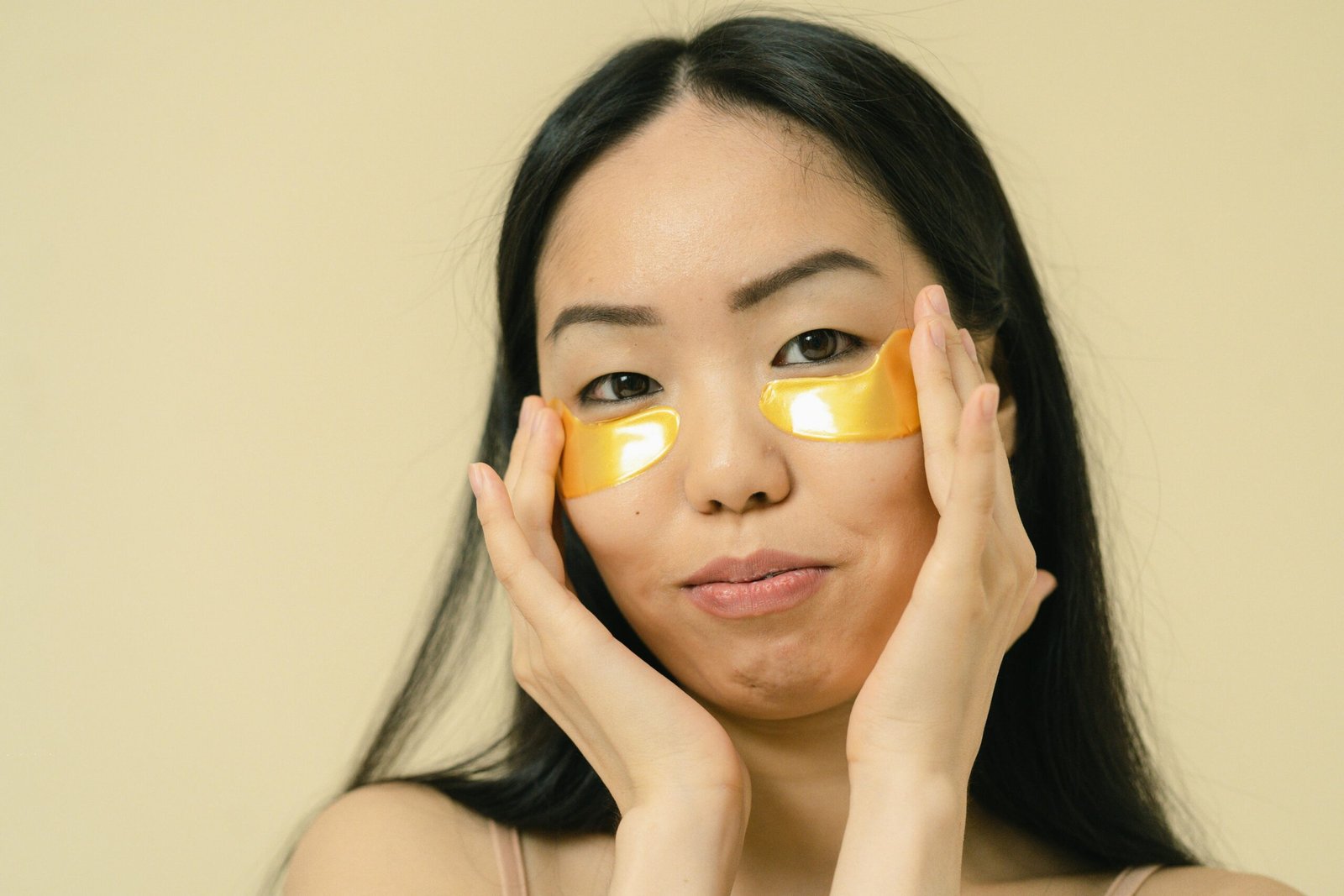 Woman with long black hair using gold eye patches for skincare in a studio.